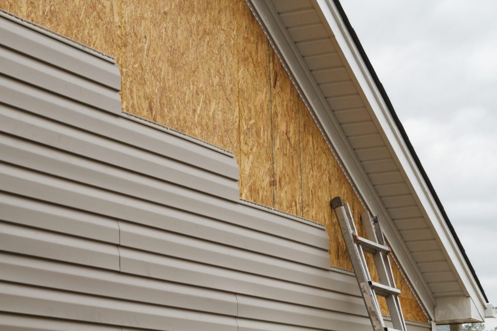 artially installed vinyl siding on a house with exposed wooden sheathing and a ladder leaning against the wall.